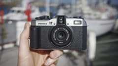 Pentax 17 compact film camera front-on, in the hand with boats in background