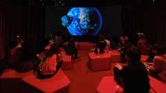 An Earth is projected on a dark screen as children sit on blocks in a dimly red-lit theater