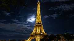 A view looking up at the Eiffel Tower lit in gold at night shining above the Paris skyline as the moon illuminates a bank of dappled clouds to the left of the steel structure.