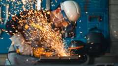A construction worker in safety helmet and gloves uses an angle grinder on a steel plate, sending a cascade of bright orange sparks across a dim industrial workshop.