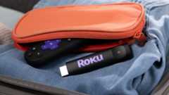 The 2025 Roku Streaming Stick and a Roku remote control emerging from an orange-colored pouch, all of which are sitting atop folded denim