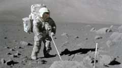 An astronaut in a white spacesuit stands on the surface of the moon with a metal detector.