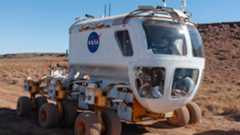 A white vehicle with six wheels and a rounded front window rolls across a dusty surface.