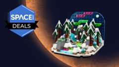 A brick-built depiction of a wintry landscape with snowy trees and running water shows the Northern Lights, all on an orange planet background.