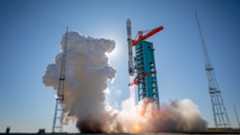A tall white rocket held up by red scaffolding begins to fire its engines, creating a cloud of white smoke. 