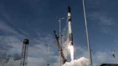 a black and white rocket lifts off above a cone of fire and smoke next two a black water tower