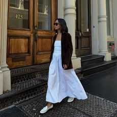 New York-based fashion influencer @linhniller on the street in SoHo, NYC, wearing a long white spring dress with flat creamy loafers and a brown suede blazer.