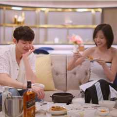 A man (Woo Sung-min) dips a spoon into a small bowl at a dining table, while a woman (Kim Go-eun) holds up a small plate while preparing to pick up food with silver chopsticks. The couple sit on a gray couch in an opulent hotel room, in front of a table holding a Korean stoneware pot, a silver ice bucket with tongs, and a bottle of Johnnie Walker bourbon. A still from 'Single's Inferno' season 5.