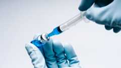 A close up shows two hands covered in blue latex gloves. The left is holding a small bottle containing blue liquid while the right hand uses a small syringe to pull out the liquid from the bottle