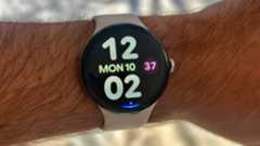 A photo of the Google Pixel Watch 4 on a man&#039;s wrist showing the Bold Utility watch face with large, colorful clock numbers and day/date info. At the display bottom is a blue Gemini Raise to Talk indicator.