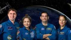 four humans stand in blue jumpsuits in front of a background of a dark earth and shadowed moon, with a small ISS in the top left. 
