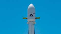 A white rocket topped with a payload fairing stands on the launch pad in a close-up set against a blue sky.