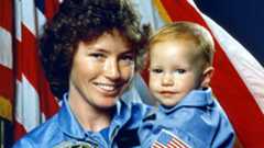 Astronaut Anna Fisher with her baby, stood in front of the American flag.