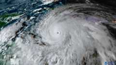 A satellite image showing a large white hurricane headed toward green land in the left side of the image contrasted with the dark blue ocean