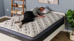 The image shows a woman wearing black dungarees lying on her side on the Helix Midnight Luxe mattress, which is placed on a white bedframe in a blue bedroom