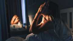 A woman sits up in bed at night, unable to sleep. She has her hands covering her face.