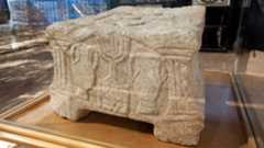a rectangular stone block carved with a menorah and amphorae sits behind glass in a museum