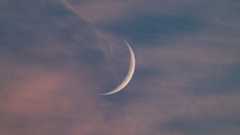 A thin crescent moon is pictured against a darkening blue sky at sunset with a sliver of its right side lit by direct sunlight. Wispy clouds are visible in the foreground.