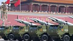 A series of green tanks holding large missiles roll on the streets below a large red building where people are standing next to China's national flags.