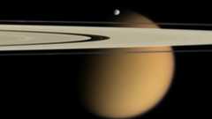 Saturn is seen as a giant yellow planet with golden rings in front and a small moon is seen as well