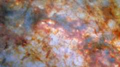 A close-in view of the centre of galaxy M82. Bright, bluish light radiating from the centre is due to stars actively forming there. A thick lane of gas, black in the centre and red around the edges, crosses the centre and blocks much of the light. Thinner strands and clumps of reddish dust cover much of the rest of the view.