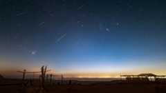shooting stars are pictured streaking through a starry night sky above a desert. A well and wooden swing set is visible in the foreground, and a glow can be seen creeping into the horizon from a settlement bordering the far side of a lake.