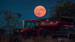 A large full moon with a red/pink hue hangs low in the sky above farming machinery in the foreground.