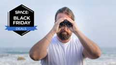 A person looking head on through binoculars with the shore and horizon in the background with a black Space.com Black Friday deals badge in the upper left corner. 
