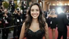  A young woman with long, dark, wavy hair is smiling broadly and looking directly at the camera while standing on a red carpet. 