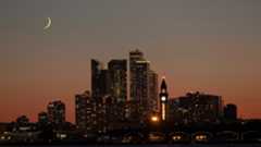 A yellow crescent moon is pictured above a city skyline. Skyscrapers are clustered in the middle of the screen with a body of water at the bottom.