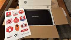 An opened cardboard box has a white box inside and a black carry bag with a sheet of stickers sitting to the left.