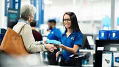 A smiling Best Buy employee wearing glasses and a blue polo shirt hands a small blue box to a customer at a checkout counter.