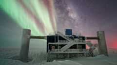 A concrete structure with large pillars and a metal staircase looms over a snowy landscape with green and red auroras illuminating the night sky 