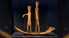 two wooden human figurines with quartz eyes and removable phalluses stand on a snake-headed boat