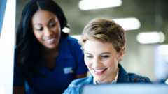 A smiling black woman in a blue uniform shirt leans in to assist a smiling white woman in a denim jacket who is looking at a tablet and a laptop screen.