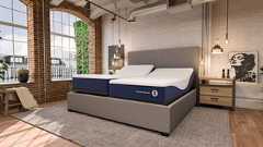 A dual-sided Sleep Number ComfortMode smart bed is shown in a modern, industrial-style bedroom with exposed brick walls and large windows. The bed features two independent mattresses on an adjustable base, with both the head and foot sections slightly elevated. The mattresses have a navy blue base with "ComfortMode" branding and a white, textured top cover. The bed is framed by a grey upholstered headboard and base, set on a grey area rug next to a wooden nightstand with minimalist decor.