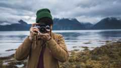 Best beginner cameras: Image shows person in woolly hat taking picture with water and mountains behind them