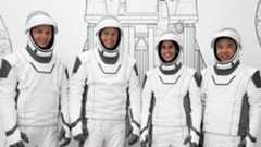 four astronauts standing in a row in spacesuits, smiling. a line drawing of a rocket is behind them