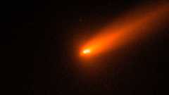 Two bright points of white light are pictured against a dark background representing the nucleus of a fractured comet, surrounded by a red hazy coma and tail streaming away to the upper right of the image.