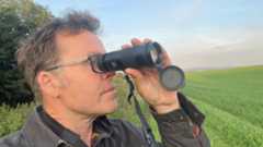 A man with glasses holding a monocular up to his right eye with a green field and blue sky in the background. 
