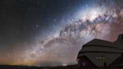 A glowing galaxy can be seen in the orange and blue night sky over the domed building of the Rubin Observatory