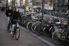 Two people cycle next to a row of parked bikes