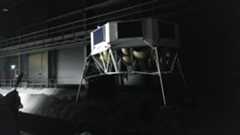 An image of a gray metal hexagonal lunar lander with four thin metal feet standing in a dimly lit warehouse with gray rocks on the floor