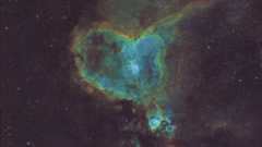 A swirling deep space nebula is pictured that takes the vague shape of a cartoonish human heart. The centre of the heart nebula is a pale blue/teal, while the outside is formed of dense dusty filaments of yellow and brown. A colorful starfield is visible in the background.