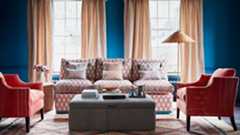 Living room with blue walls, patterned red sofa, armchair, striped ottoman and rattan floor lamp