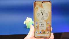 Google Pixel 10 Pro XL in Porcelain against a blue background with a green neon Android figurine in the background