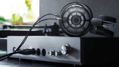 Product photography of the Audio-Technica ATH-ADX7000 over-ear headphones atop a headphone amp with a window in the background