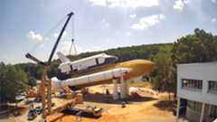 a crane lifts a full-size model of a nasa space shuttle onto its mock orange fuel tank and white solid rocket boosters in a horizontal position, with trees in the background