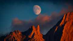 A near full moon is pictured in a blue sky above the jagged peaks of a mountain range. Both the mountains and the clouds below the moon are lit by the golden light of dusk.
