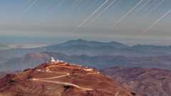 A large mountain is seen with a glowing windy road leading up it to two buildings at the top, with the rest of the rocky desert landscape all around and red streaks in the sky above.